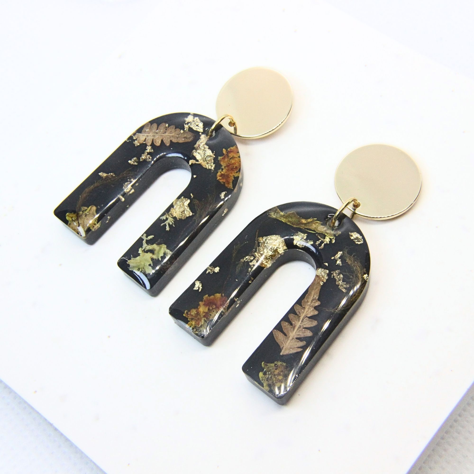 Black resin arch earrings with grass seed inlay and gold flecks, finished with gold plated sterling silver disc tops.