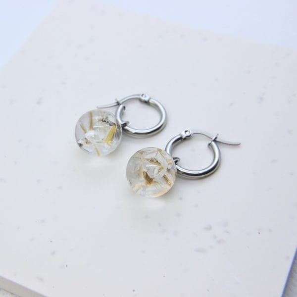 Clear resin globe hoop earring with wild daisy seeds preserved inside and stainless steel hoop fitting.
